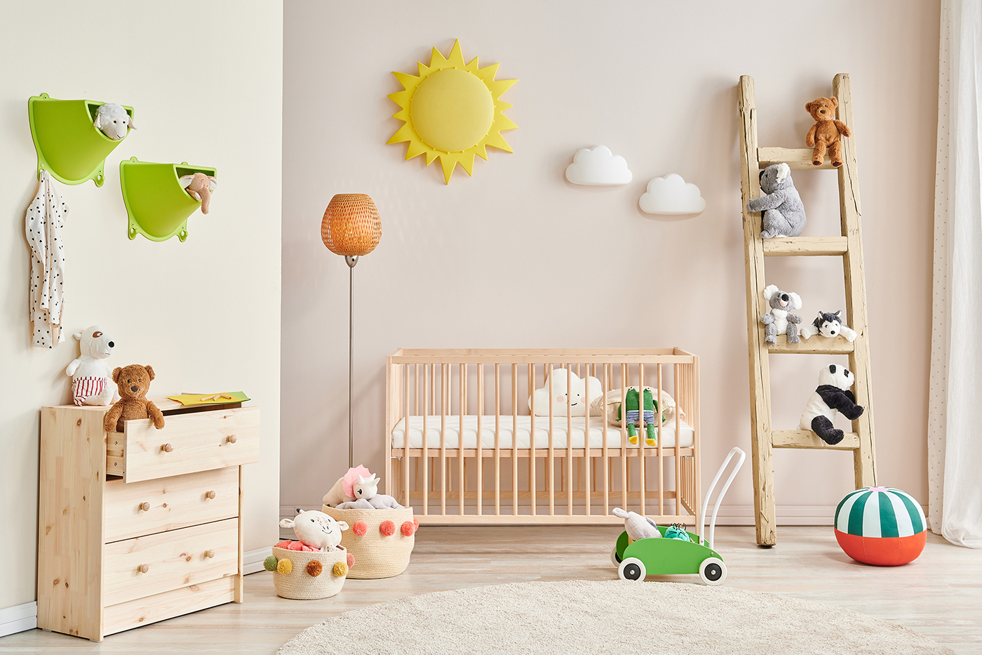 Light-wood crib and dresser in pastel nursery; ladder shelves hold plush toys, sun and cloud wall decor, green push toy on rug.