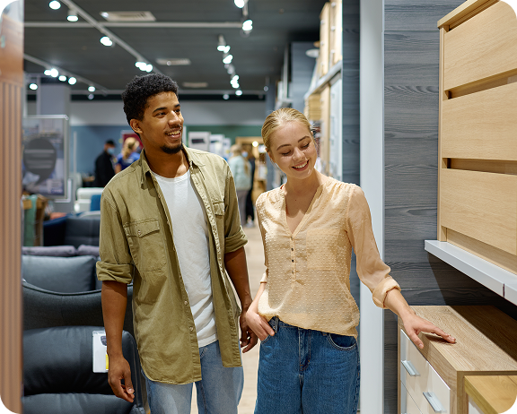 A smiling couple examines modern wooden cabinets in a furniture store, surrounded by various home furnishings.