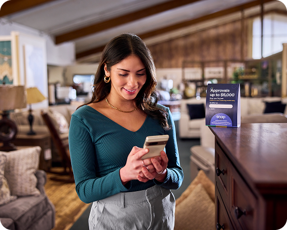 Woman using smartphone in a furniture store, standing near a sign about approval up to $5,000.