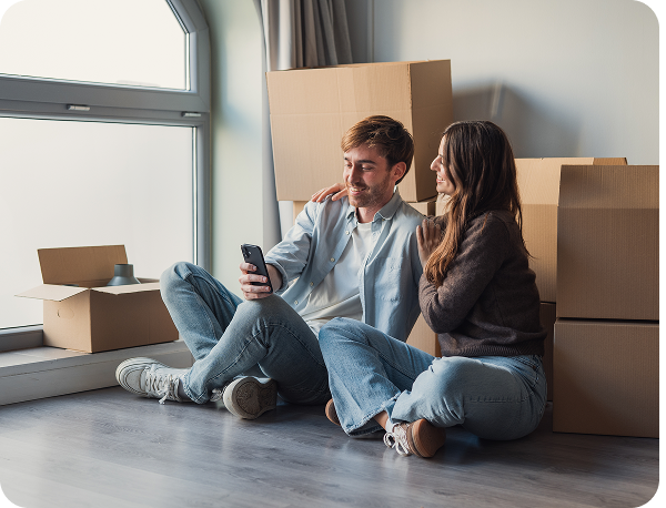 A couple sits on the floor of a new home, surrounded by moving boxes, smiling at a smartphone.