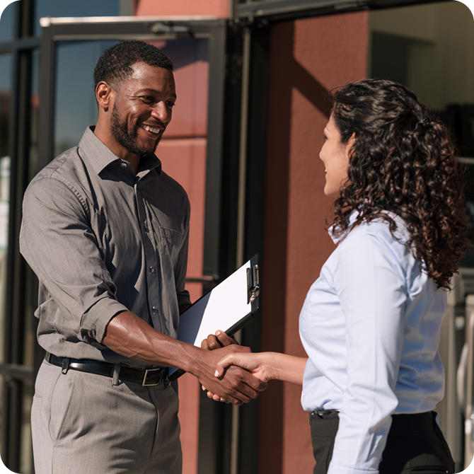A man and woman smiling and shaking hands outside a building. The man holds a clipboard.
