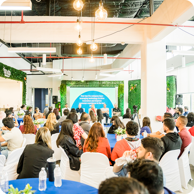 A group of people seated in a bright, modern conference room, attentively listening to a panel discussion in front of a large screen.