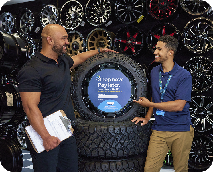 Two men smiling by a tire display in a shop, standing near a "Shop now. Pay later." sign with various wheels in the background.