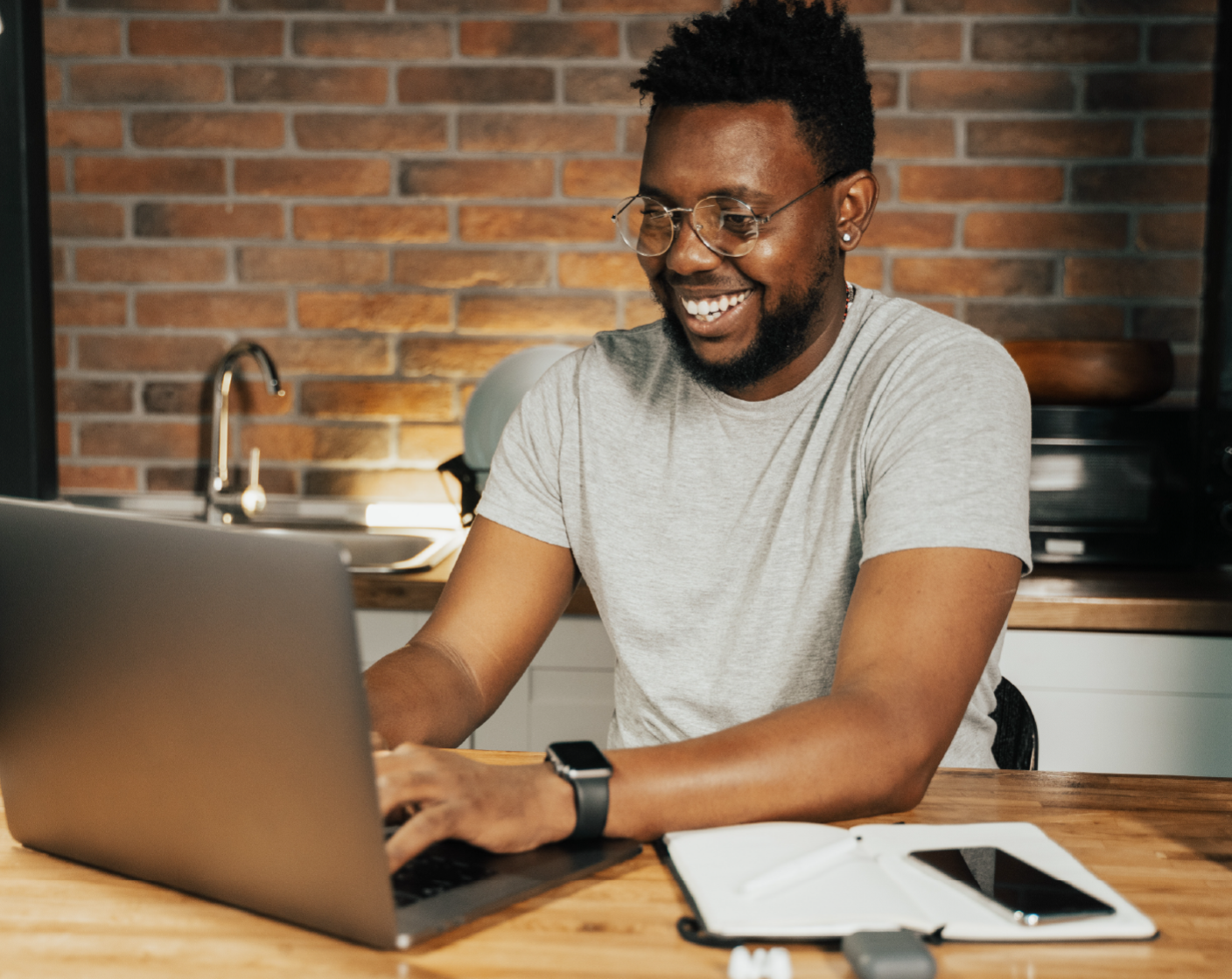 Cheerful man with glasses working on laptop in home kitchen, brick wall background