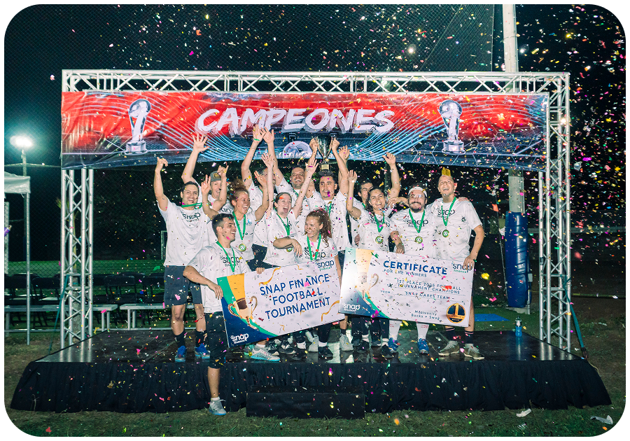 A soccer team celebrates on a podium, holding a "Champions" banner, certificates, and a trophy. Confetti fills the air under a "Campeones" sign.