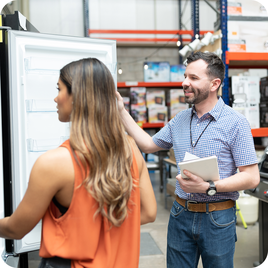 A salesperson shows a fridge to a customer in a store, holding a notepad. Shelves and product displays are in the background.