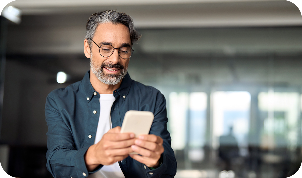 A man with glasses and gray hair smiles while looking at his phone in a modern, bright office setting.