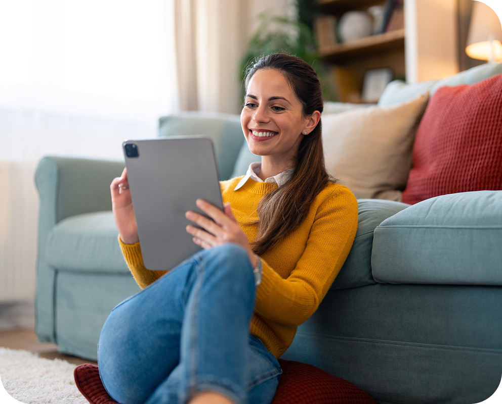 Smiling woman in a yellow sweater sitting on the floor, leaning against a sofa, using a tablet in a cozy living room.