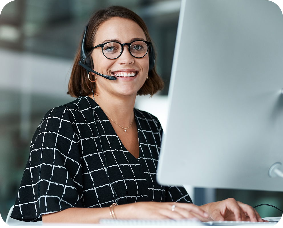 A woman with glasses and a headset smiles while working on a computer in an office environment.