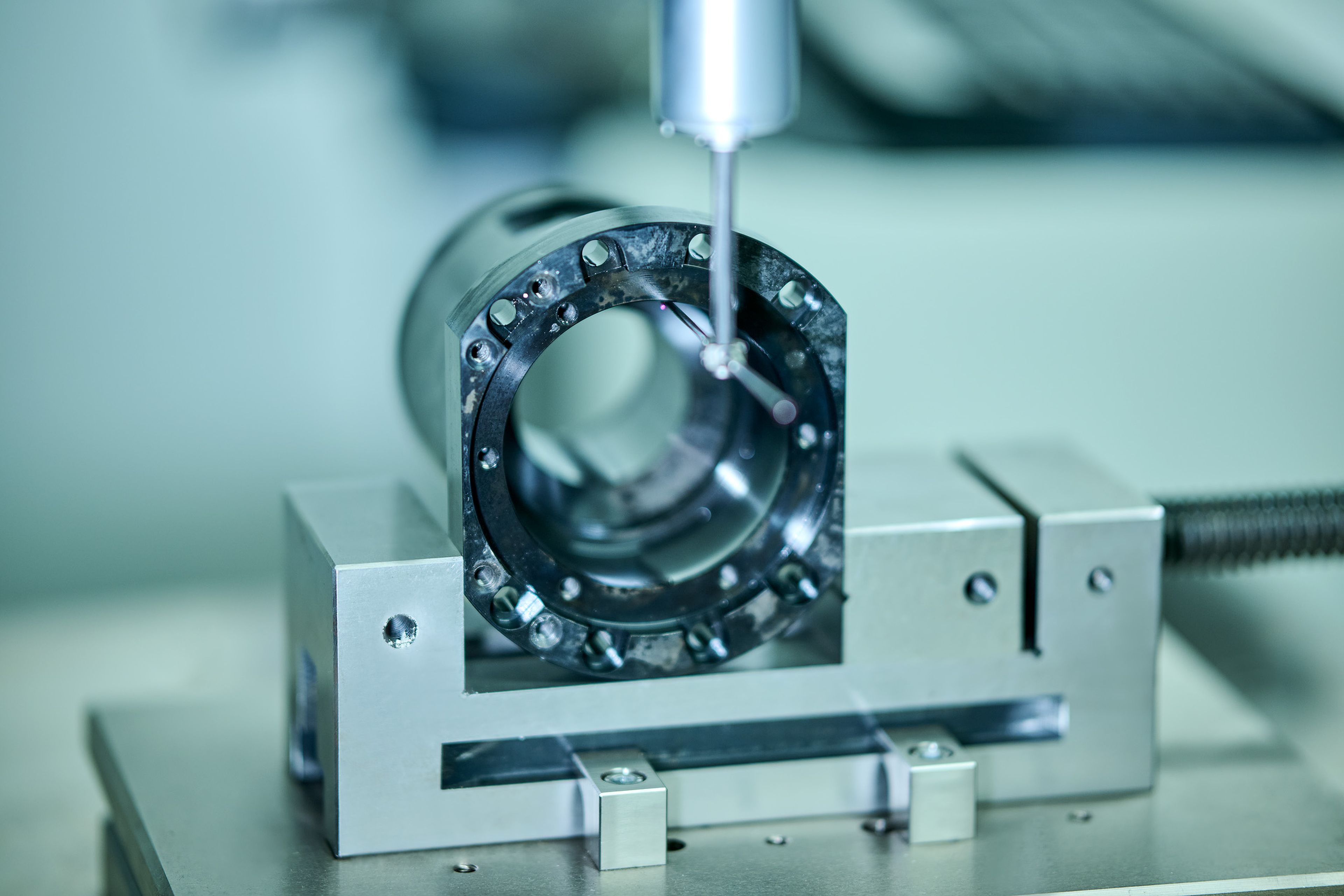 Nahaufnahme eines Präzisionsmessgeräts, das ein zylindrisches Metallteil in einem Schraubstock in einer Laborumgebung prüft.