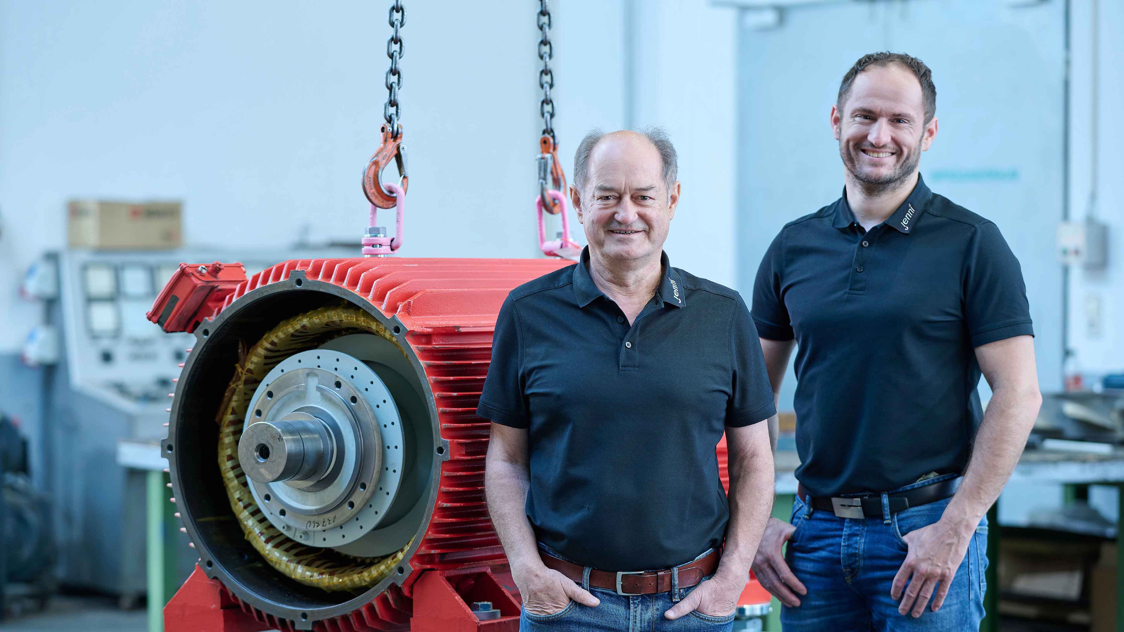 Zwei Männer in schwarzen Hemden stehen in einer Werkstatt neben einer großen roten Industriemaschine, wobei ein Mann lächelt und der andere in die Kamera schaut.
