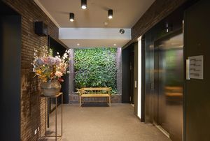Binnenruimte met bakstenen muren, een bloemstuk op een standaard, liften aan de rechterkant en een houten bank voor een beplante muur.