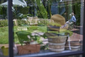 De weerspiegeling van binnenstoelen in het raam geeft een zonnig terras in de tuin weer, waar een oudere persoon in een rolstoel zit, vlakbij bankjes en planten.