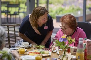 Een vrouw zit in een zonnige ruimte aan een gedekte tafel. Een vrouw staat naast haar, buigt voorover en kijkt haar aan.