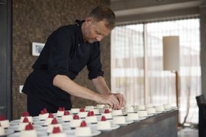 Een man in zwart koksuniform staat in een keuken gebogen over een aanrecht vol schoteltjes met panna cotta. Hij doet er framboosjes op.