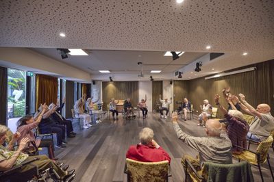 Ruime activiteitenruimte met ouderen op stoelen die in een cirkel op een houten vloer staan. Ze hebben allemaal beide armen omhoog.