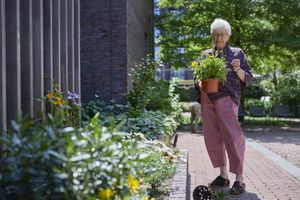 Een oudere dame met kleurrijke kleding staat met een plant in haar handen buiten naast een bloemenperk.