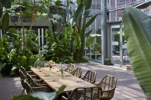 Zonovergoten ruimte met glazen deuren, lange houten eettafel en rieten stoelen, omgeven door grote tropische planten.