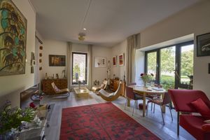 Zonnige, moderne woonkamer met rood tapijt, gebogen houten ligstoelen, eettafel bij groot raam en kunstwerken aan de muren.