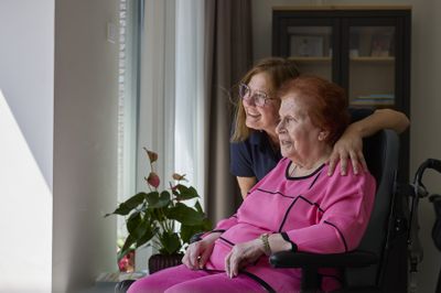 Een mevrouw met felroze kleding in een rolstoel en een mevrouw met bril kijken samen door het raam naar buiten.