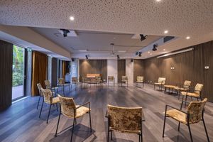 Ruime activiteitenruimte met stoelen die in een cirkel op een houten vloer staan, grote ramen met gordijnen en met hout beklede wanden.