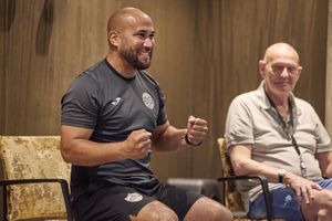 Een man in sportkleding zit in actieve houding met gebalde vuisten in een stoel naar een oudere man.