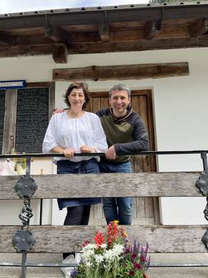 A woman and man smiling on a wooden balcony with colorful flowers in front, standing in front of a white building with a wooden door.