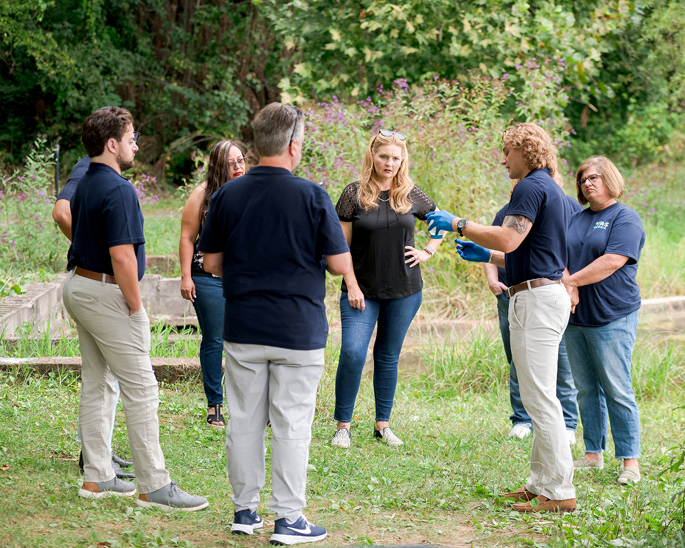A group of people stand in a circle outdoors, listening while one of them speaks. They are surrounded by greenery and appear focused..