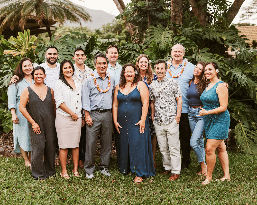 A group of people smiling and posing outdoors, wearing leis and casual attire, surrounded by lush greenery and trees.