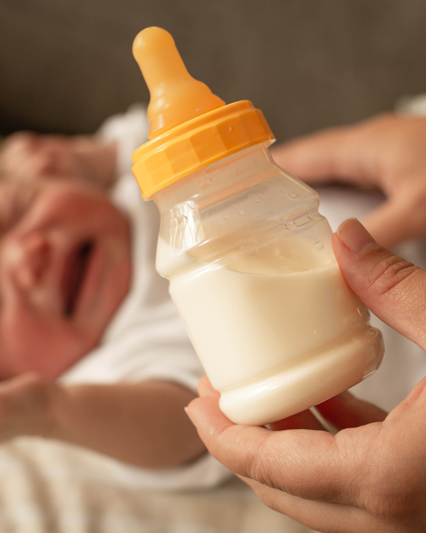 Close-up of hands holding a baby bottle with milk, while a crying baby lies in the background.