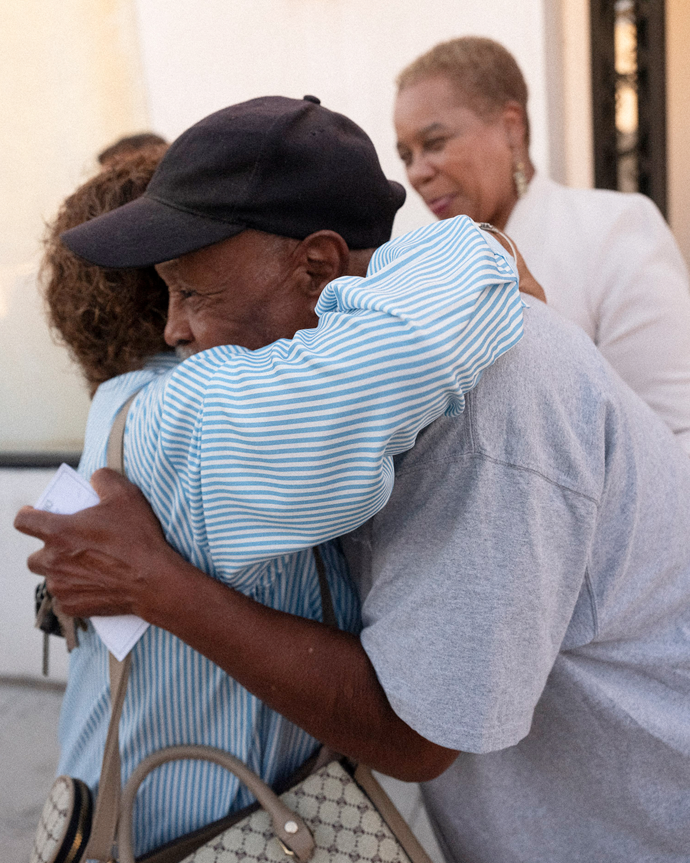 Two people hug. A third person stands nearby, smiling.