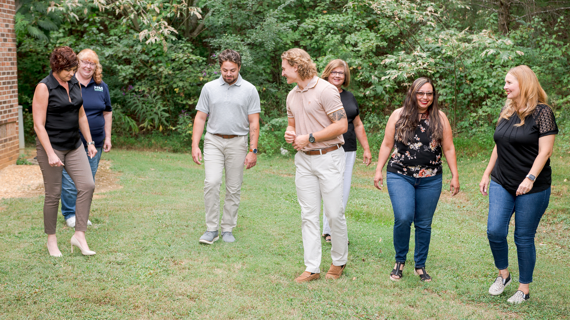 A group of seven people walking and smiling on a grassy area, surrounded by trees.