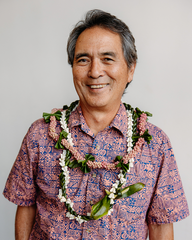 Smiling man wearing a patterned shirt and multiple floral leis against a plain background.