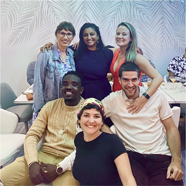 A group of six people smiling and posing together with a patterned wall in the background.