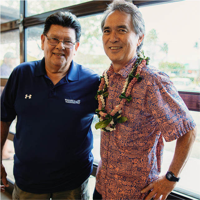 Two men smiling indoors; one wears a blue polo, the other a patterned shirt with a lei. They're standing in front of large windows.