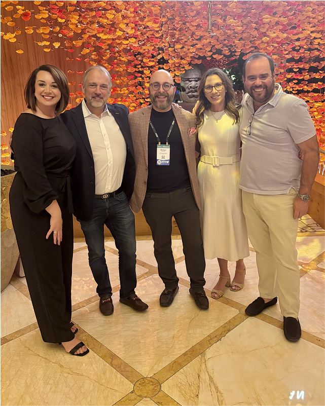 Five people smiling and posing together in front of a decorative wall with orange leaves.
