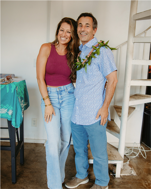 A smiling woman in a maroon top and jeans stands next to a man in a floral shirt with a lei, inside a room with a spiral staircase.