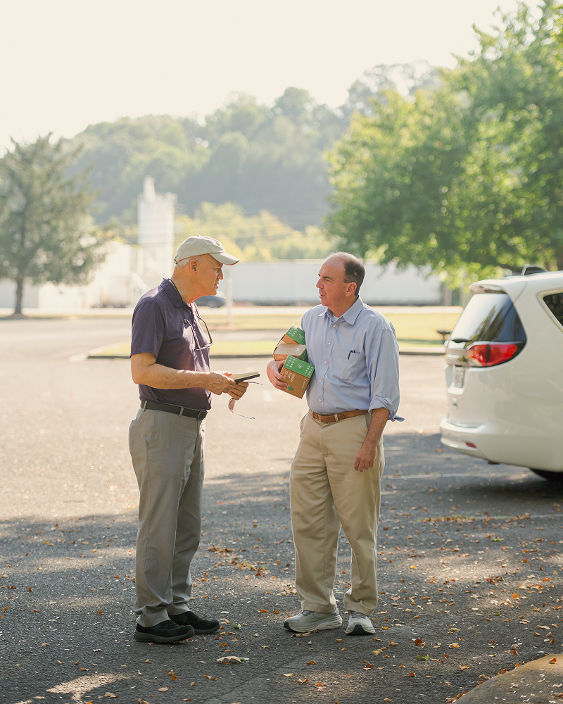 Two men stand in a sunny parking lot, talking. One holds several boxes. Trees and a parked car are in the background.