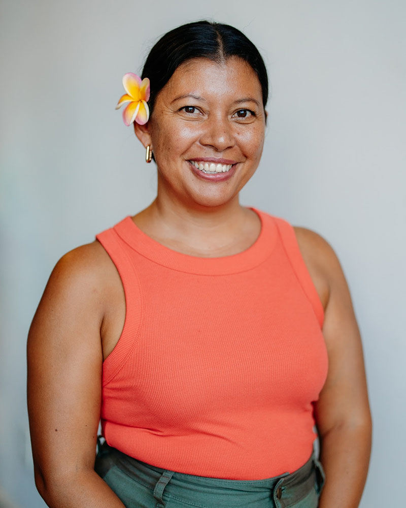 Smiling woman in an orange tank top with a flower in her hair, standing against a plain background.