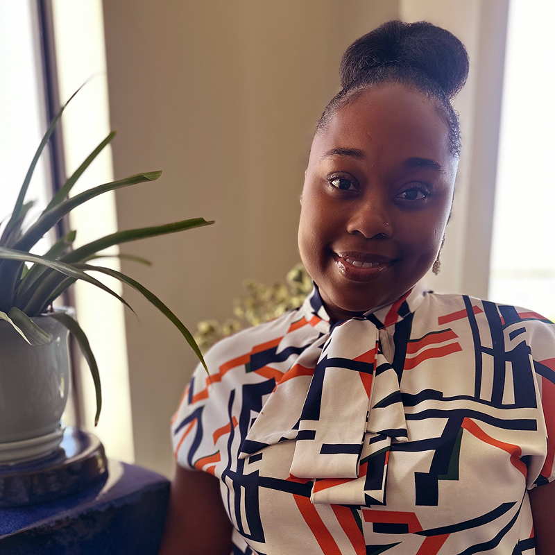 Smiling woman with a bun hairstyle wearing a patterned blouse, stands beside a potted plant in a well-lit room.