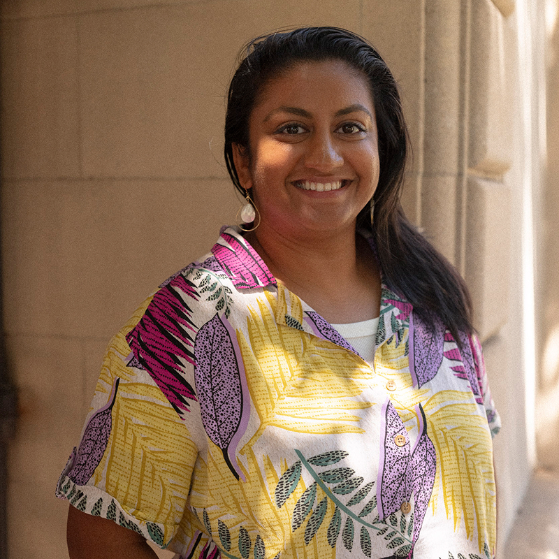 Smiling person with long hair in a colorful, patterned shirt stands by a stone wall, partially in sunlight.