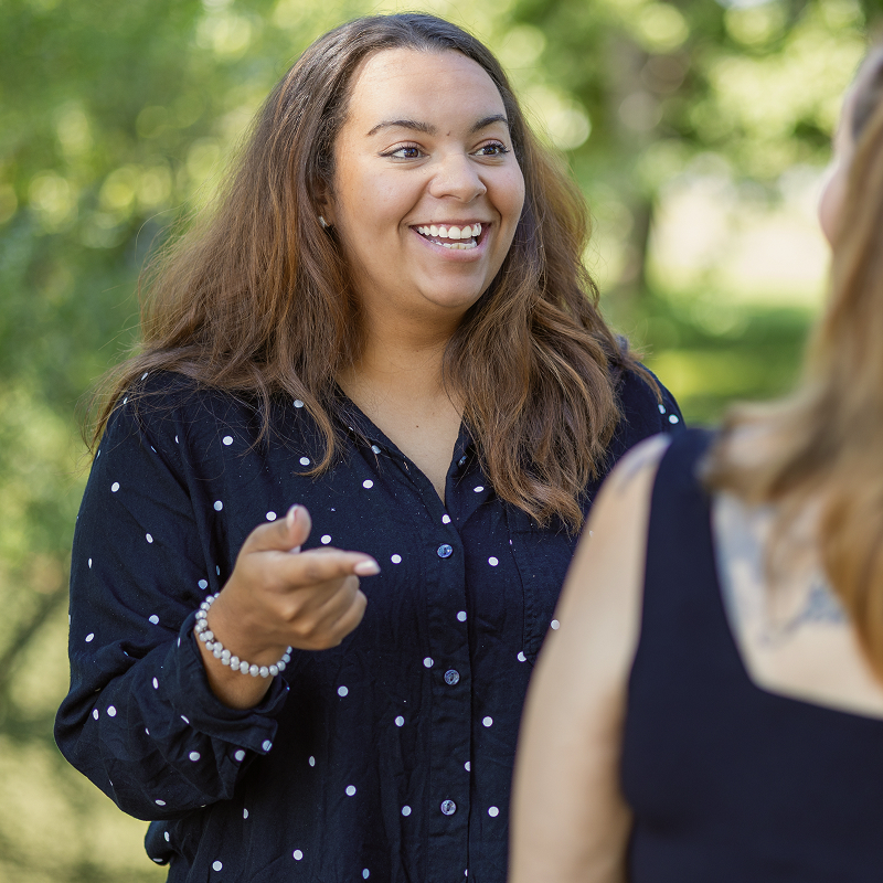 Smiling woman in a polka dot shirt gestures while talking to another person, with green foliage in the background.
