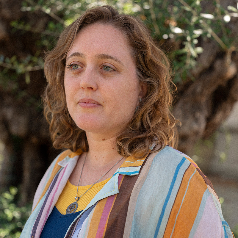 Woman with wavy hair wearing a striped shirt, standing outdoors with a tree in the background.