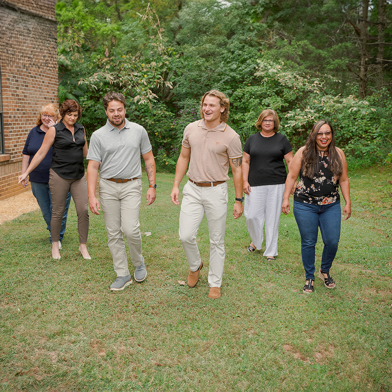 A group of six people walking on grass, smiling and talking, with trees and a brick building in the background.