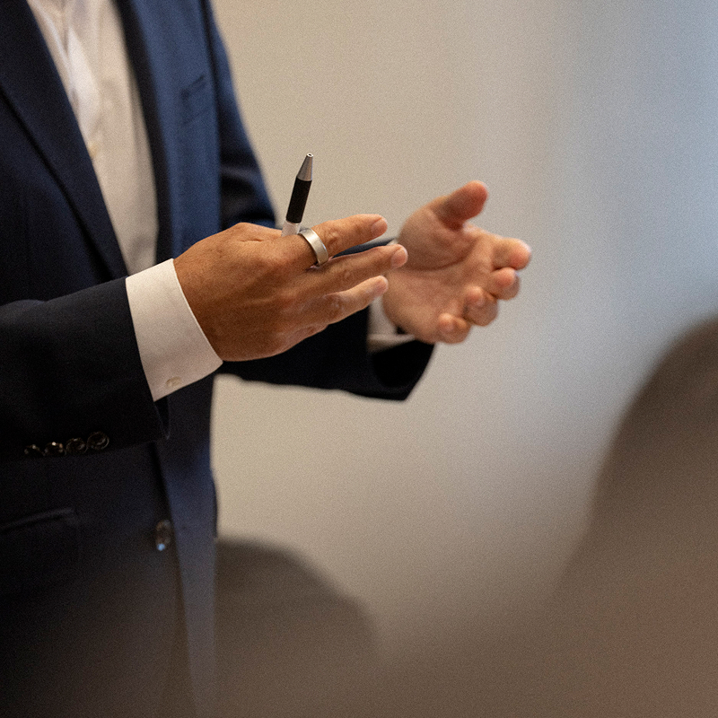 A person in a suit gestures while holding a pen.