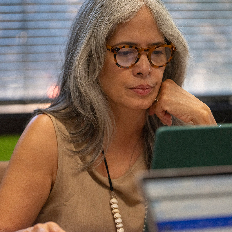 A woman with long gray hair and glasses focuses intently on a laptop, seated in an office with blinds in the background.