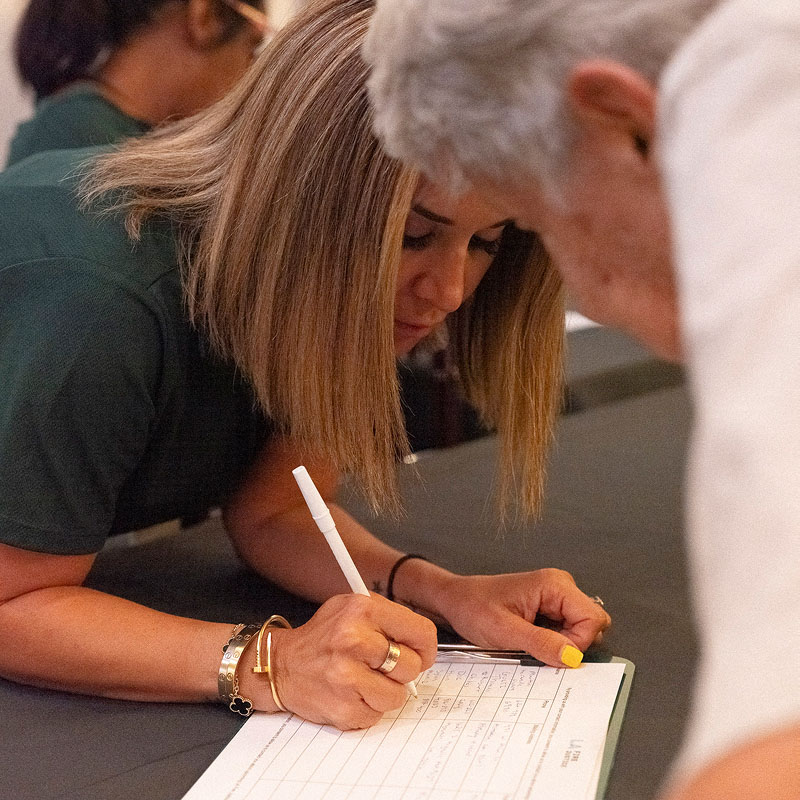 A woman fills out a form, while an older woman watches from across the table.