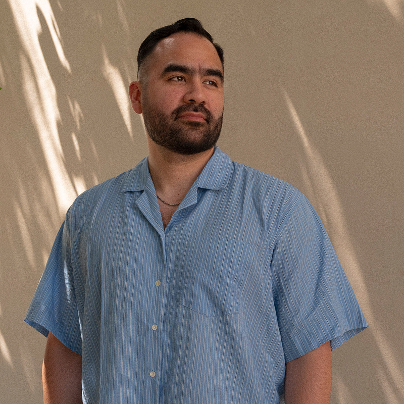 Man with short hair and beard in a blue striped shirt stands against a sunlit wall with leaf shadows.