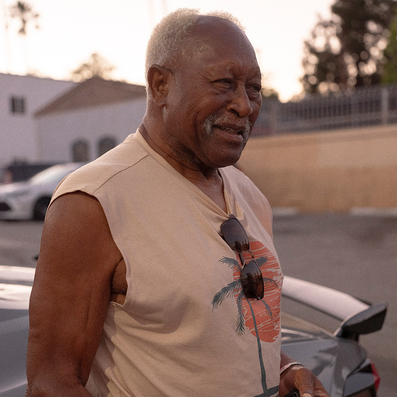 Elderly man smiling outdoors, wearing a sleeveless shirt with a palm tree design.