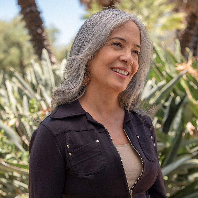 A smiling woman with long gray hair stands outdoors in front of green foliage and palm trees, wearing a dark jacket.